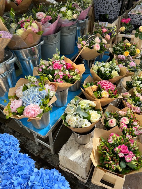 A vibrant display of fresh floral bouquets arranged in metal buckets and wrapped in brown craft paper, featuring a variety of flowers in soft pinks, whites, blues, and yellows. The arrangements includ
