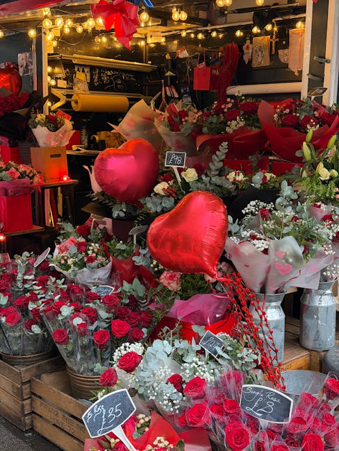 An assortment of floral arrangements displayed at a florist shop with warm hanging string lights overhead. The display features numerous bouquets predominantly composed of red roses, complemented by w