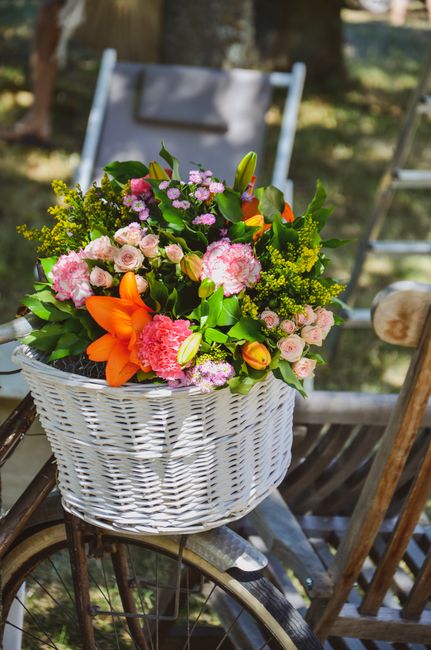 A vibrant floral arrangement in a white wicker basket featuring orange lilies, soft pink carnations, small light pink roses, green hypericum berries, and lush greenery. The basket is placed on a vinta
