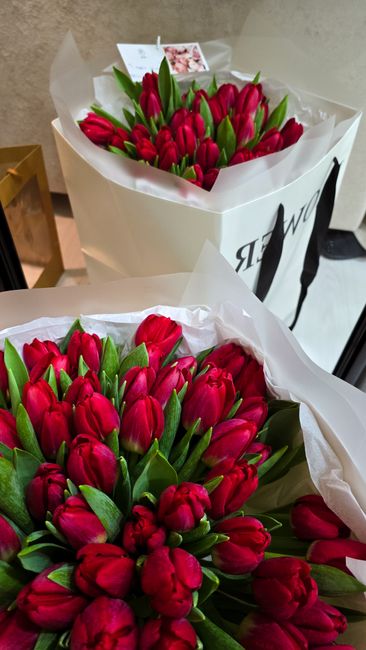 Two bouquets of vibrant red tulips with green leaves arranged closely together, wrapped in white floral paper, placed in front of a white gift bag with black handles and the logo of floristtwickenham.