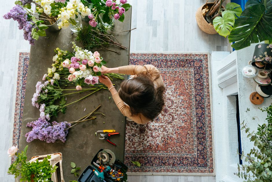 A top-view image of a florist arranging a floral bouquet on a grey table. The arrangement includes various fresh flowers such as pink and white roses, purple lilacs, and additional greenery. The flori