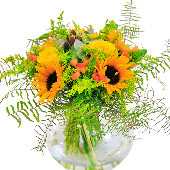 First impressions of this bouquet are woven with light and energy-the bright faces of sunflowers and rich, sunshine-yellow roses interlaced with lush, textured greenery. Each sunflower radiates a sense of joy found along Twickenham's bustling streets, its chocolate-brown core offsetting a halo of golden petals that nearly glimmer in the imagined sunlight. Yellow garden roses, boastful yet elegant, unfold their tightly layered petals alongside playful tufts of delicate golden filler, reminiscent of summer wildflowers at Radnor Gardens. Ruby red Hypericum berries nestle within the foliage, their gloss contrasting against the silken petals, while slender spikes and feathery ferns spill gently outwards, conjuring motion and a touch of the wild. Subtle eucalyptus leaves add a whisper of blue-green, both in scent and tone, rounding out the bouquet's lively palette. The spherical vase, clear as the Thames on a crisp morning, reveals a tangle of fresh stems and water below, reinforcing the bouquet's freshness and artisanal craftsmanship from a florist near Twickenham's heart. Perfect for sharing congratulations, marking birthdays, or simply illuminating a home, this carefully composed arrangement whispers of open spaces and shared celebrations beneath the local skyline.